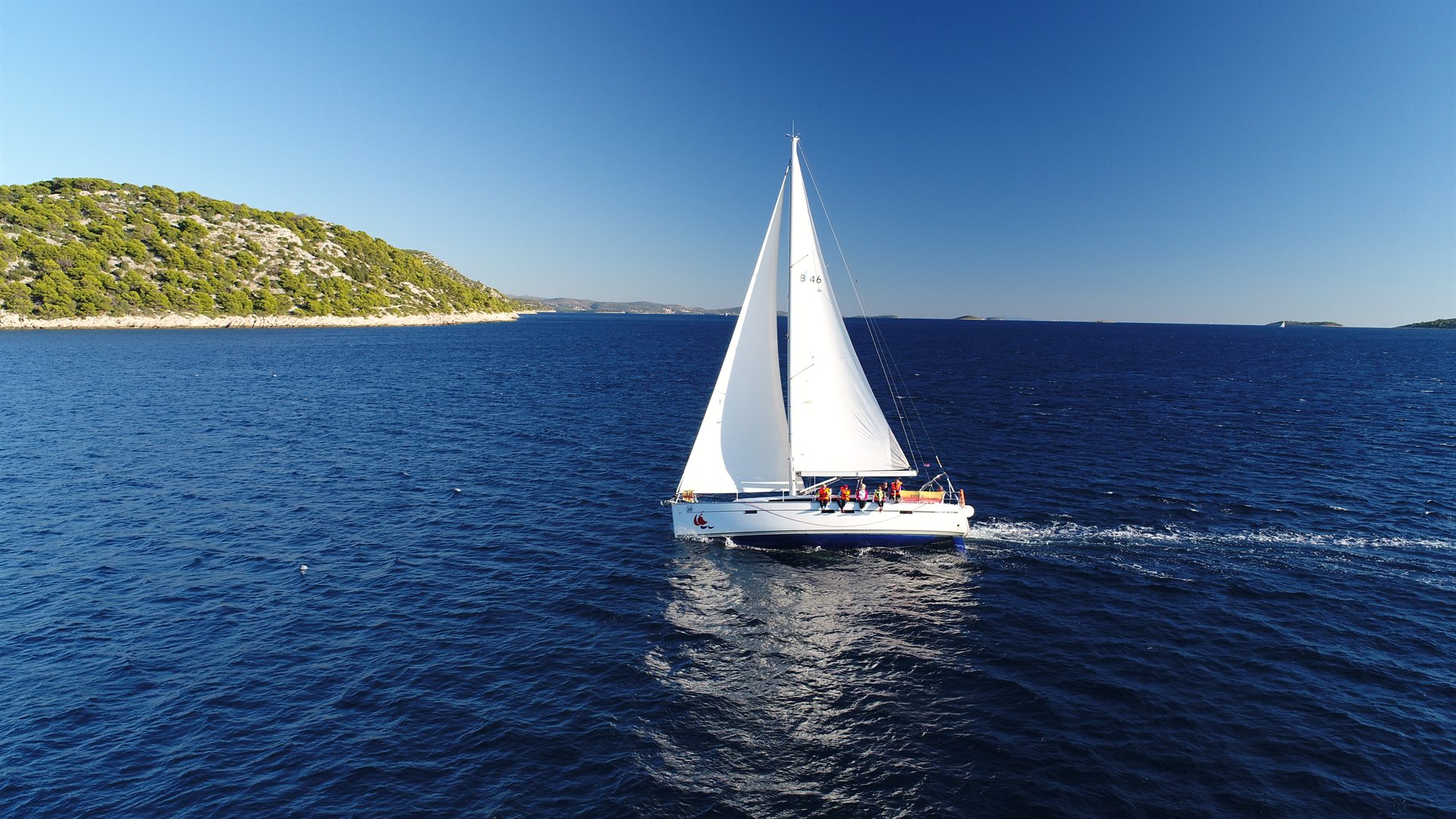 Biały jacht z pełnymi żaglami i pasażerami w kamizelkach na tle ciemnoniebieskiego morza i zielonej wyspy