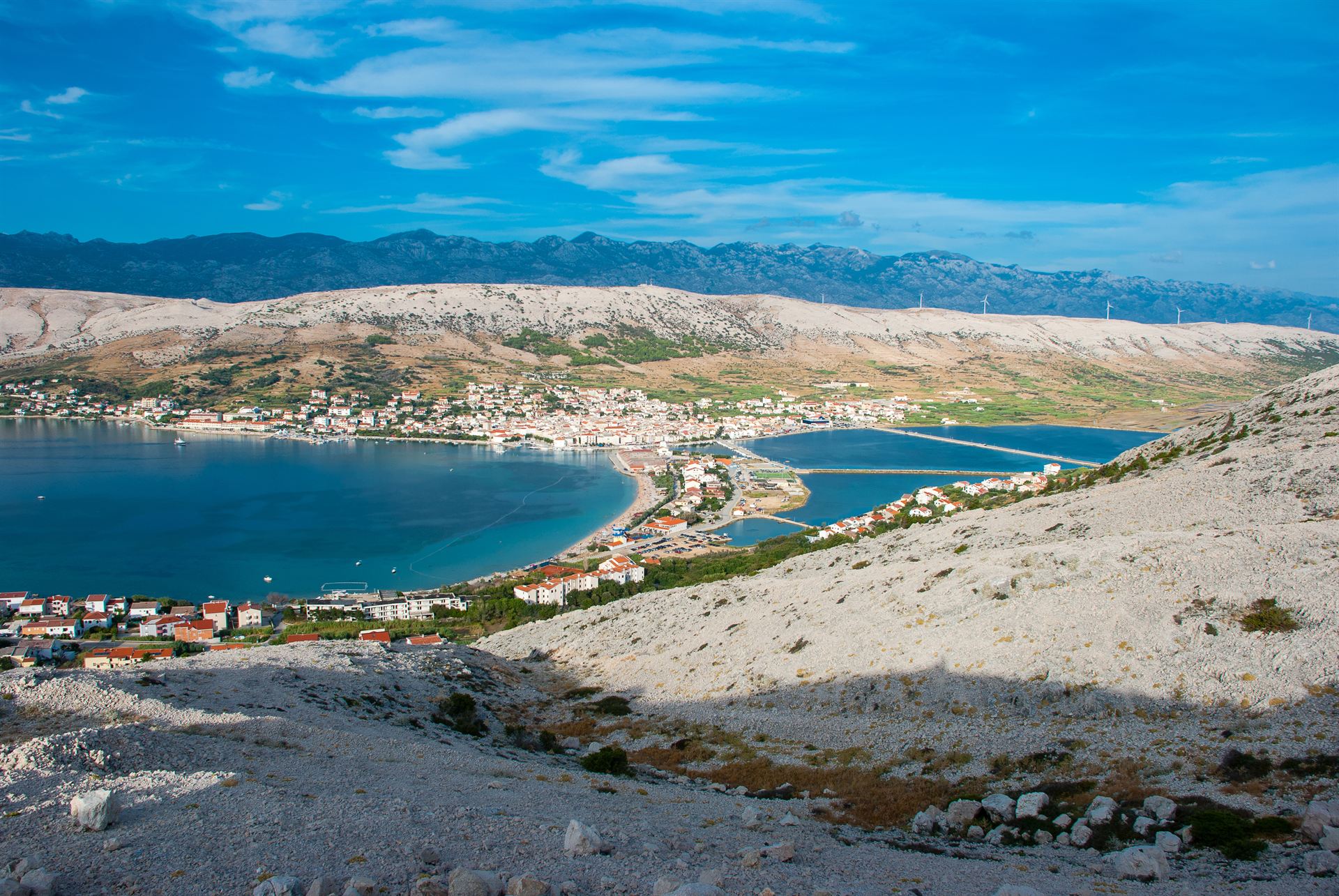 Nadmorska miejscowość na wąskim półwyspie z czerwonawymi dachami, turkusową zatoką i górskim pasmem w tle