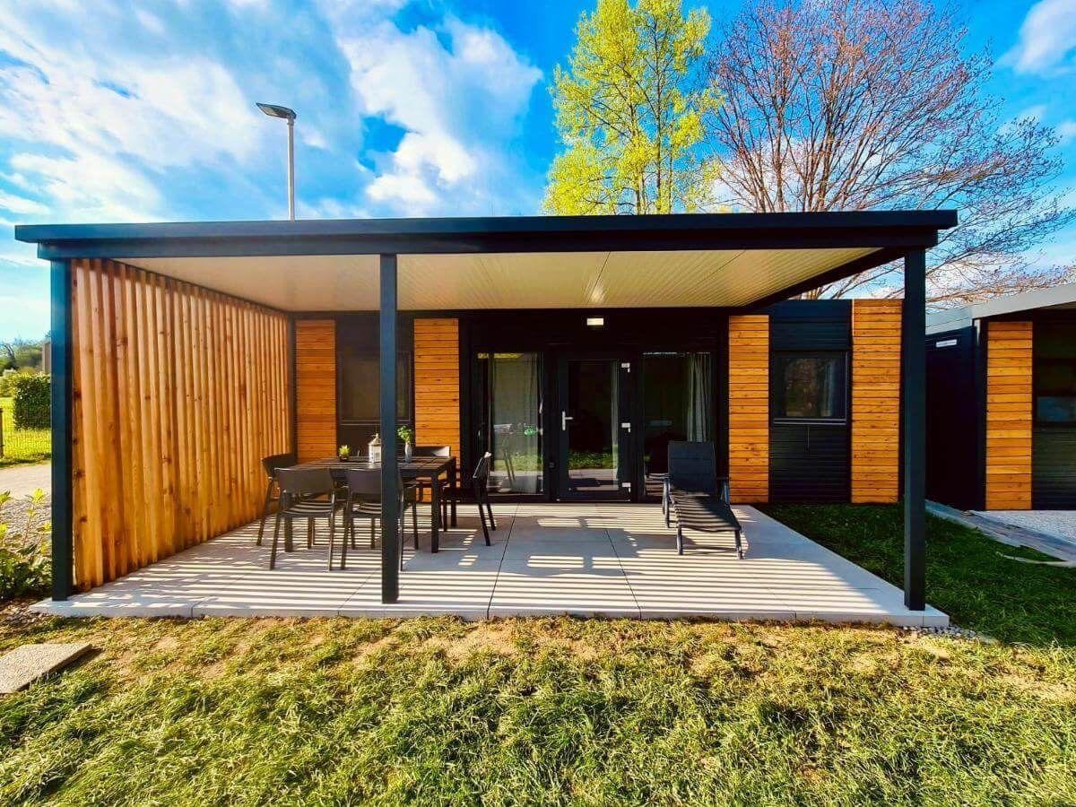 Nowoczesny drewniany domek z zadaszonym tarasem, czarną ramą, stołem z krzesłami i leżakiem na trawie