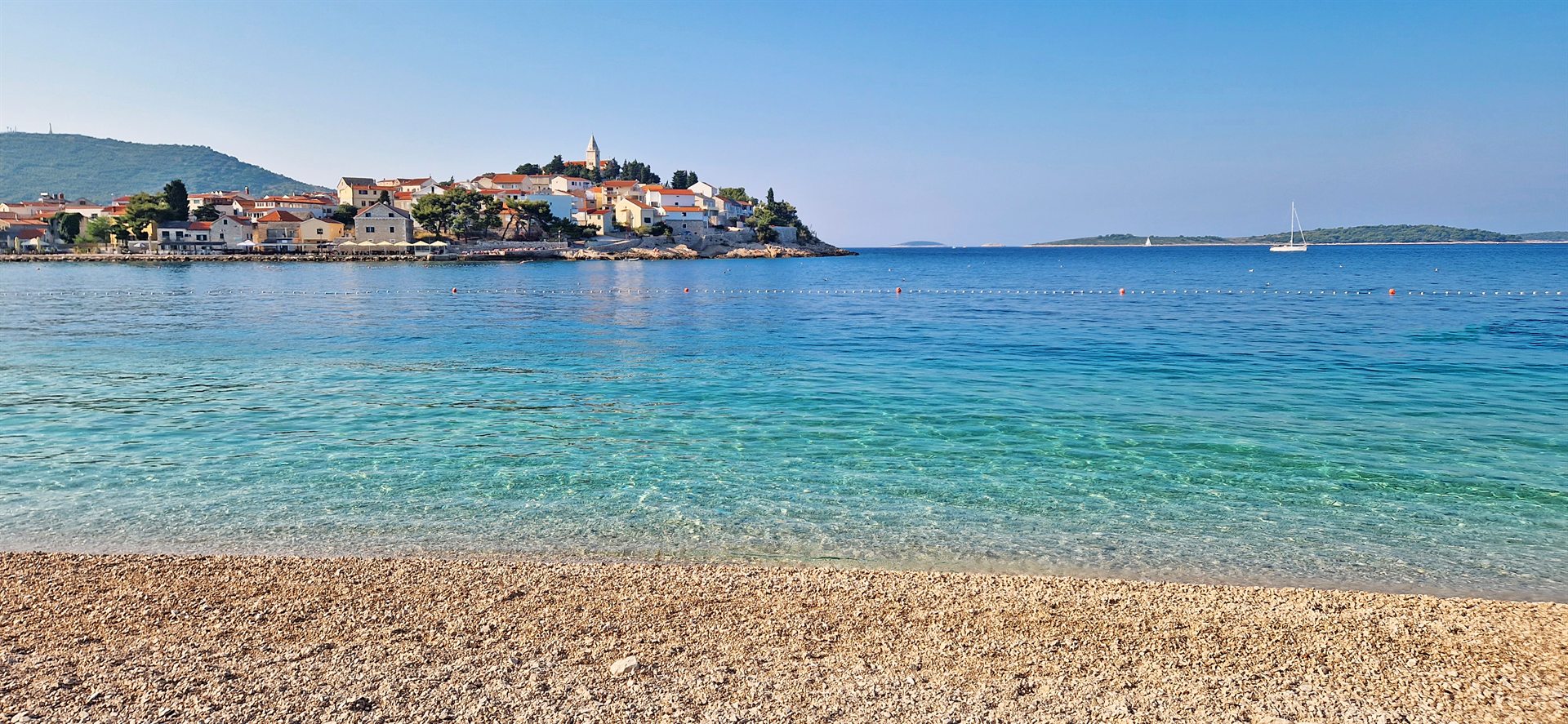 Kamienista plaża, przejrzysta turkusowa woda i nadbrzeżne miasteczko z czerwonymi dachami i wysoką wieżą kościelną